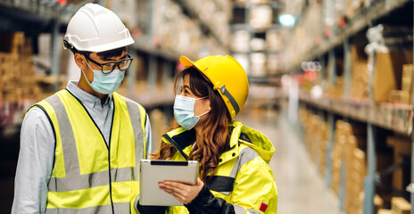 Asian engineer man and woman in helmets in quarantine for coronavirus wearing protective mask working in new normal at shelves with goods background in warehouse.logistic and business export