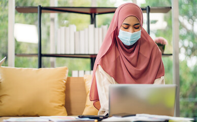 Young asian muslim woman relaxing using laptop computer working and video conference meeting online chat in quarantine for coronavirus wearing protective mask with social distancing at home