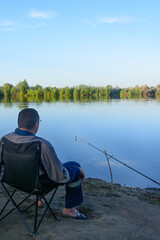 Fisherman sits on bank of river and looks at fishing rod with bells. Morning fishing. Selective focus, blur