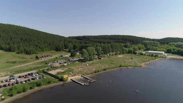 Aerial view of city beach on the outskirts of a small provincial town. People lie on the grass, swim in a pond, relax, dive, ride a jetski. Near forest, mountain and parking with many cars. Summer sun