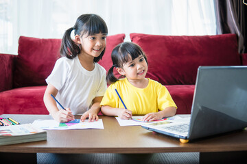Two asian child girl students study online with teacher by video call together. Siblings are homeschooling with computer laptop during quarantine due to Covid 19 pandemic.