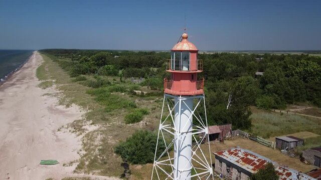 Beautiful aerial view of white painted steel lighthouse with red top located in Pape, Latvia at Baltic sea coastline in sunny summer day, wide angle ascending drone shot moving forward close