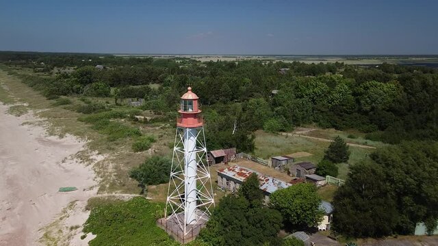 Beautiful aerial view of white painted steel lighthouse with red top located in Pape, Latvia at Baltic sea coastline in sunny summer day, wide angle descending drone shot moving forward