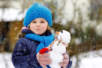 Cute little toddler girl making mini snowman and eating carrot nose. Adorable healthy happy child playing and having fun with snow, outdoors on cold day. Active leisure with children in winter