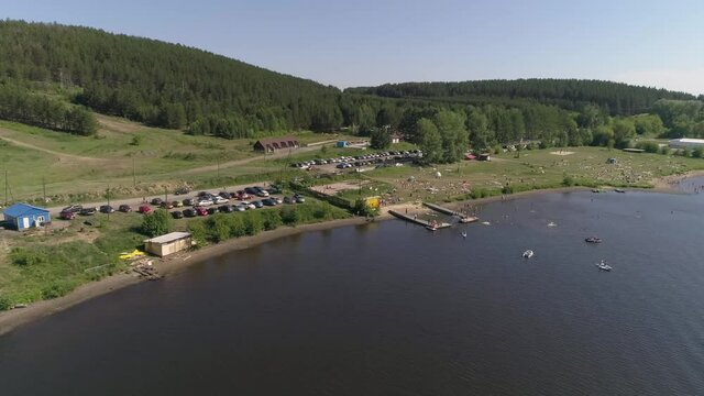 Aerial view of Summer vacation at the city beach in small provincial town. People lie on the grass, swim in a pond, relax, dive, people are boating, sunbathing. Near forest and parking with many cars.