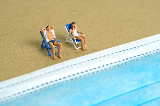 Miniature people toy figure photography. Safety travel concept, Men and girl relaxing at beach chair in front of blue face mask