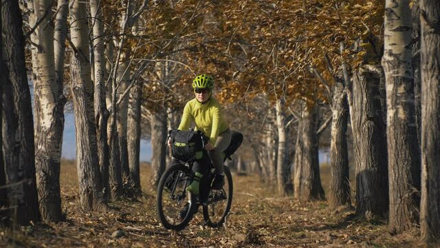 The woman travel on mixed terrain cycle touring with bikepacking. The traveler journey with bicycle bags. Sportswear in green black colors. The trip in magical autumn forest, arch, alley, avenue.