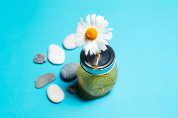 Close-up of chamomile flower in straw of juice glass mug on blue background near stones.