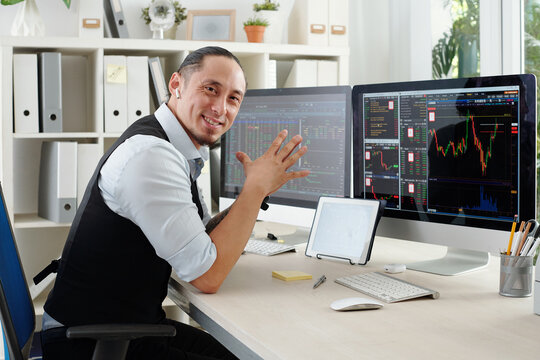 Portrait of cheerful trader sitting at his office desk with various charts and stock market data on computer screen