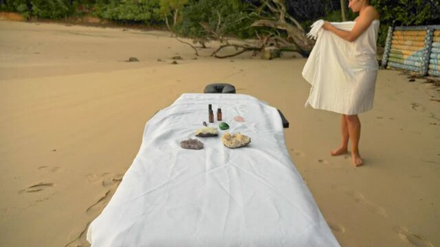 Yoni Eggs And Essential Oils On Massage Table At Echo Beach. Female Client Standing On Sandy Shore. tilt-up