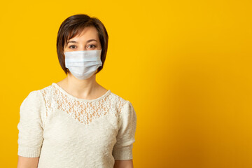 Lovely dark haired lady has epidemic disease, wears protective medical mask, poses against yellow background, has infection, stands indoor. Health care and people concept.