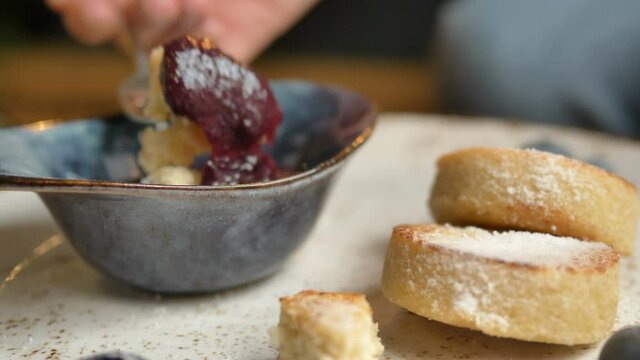 Cheesecake piece dipping in blue sauce bowl with delicious bright purple berry jam in vegan cafe extreme closeup slow motion