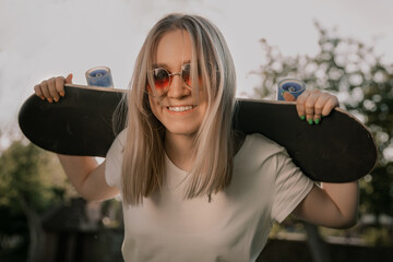 Young blonde girl holding her skateboarde, looking at the camera and smiling.
