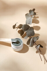 beauty, cosmetics and object concept - blue cosmetic clay mask in jar with wooden spatula and branch of eucalyptus populus on beige background