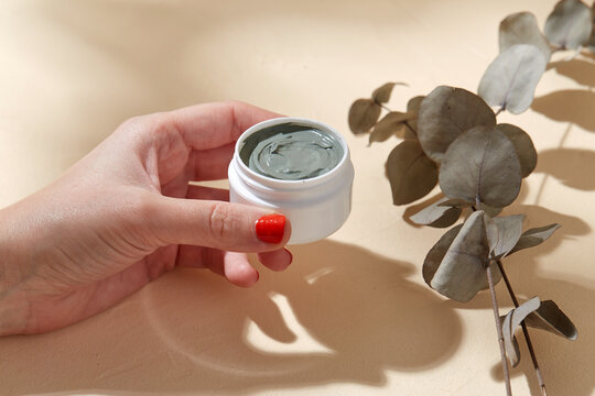 Beauty, Cosmetics And Bodycare Concept - Hand Holding Jar Of Blue Cosmetic Clay Mask Over Branch Of Eucalyptus Populus On Beige Background