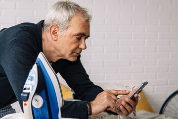 Mature man using mobile phone while ironing shirt at home