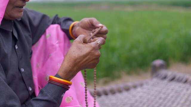 Close up shot of an aged woman  praying while using beads - Religious practice in Hindu culture. A traditional old lady using wooden beads during her prayers while she is near an agricultural land ...