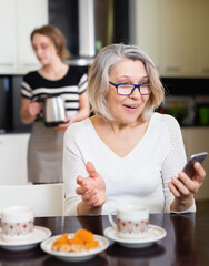 Mature woman absorbed in her smartphone while adult daughter making tea in home kitchen