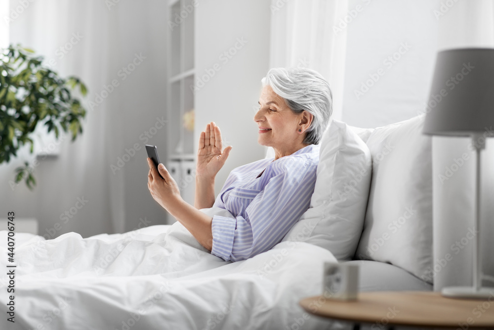 Wall mural old age, technology and people concept - happy smiling senior woman in pajamas using smartphone sitting in bed and having video call at home bedroom