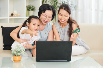 Cheerful little girl asking mother and grandmother to buy her toy or book at online store when they...