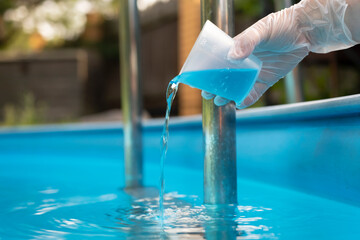 Hand holding and pouring algicide blue liquid from glass into water of swimming pool. Water...