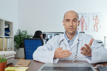 General practitioner looking at camera, gesturing and explaining new methods to treat bacterial pneumonia, his colleague checking patient x-rays in background
