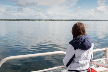 Teenager girl looking at the ocean from a deck of a ferry ship. Galway bay, Ireland. The model back to viewer. Blue cloudy sky. Girl with long hair on ocean voyage enjoys scenery. Travel concept