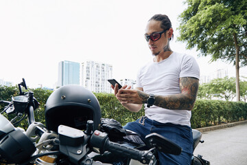 Fit handsome man in sunglasses sitting on motorcycle and reading text messages or notifications on smartphone