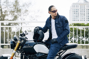 Stylish man in sunglasses, jeans and denim jacket sitting on motorcycle, taking off helmet and...