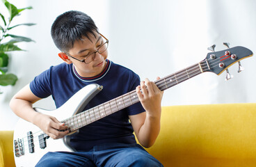 Portrait shot of handsome smiling young boy with eyeglasses enjoying playing the bass guitar. Junior bassist in casual clothes sitting on couch and holding an instrument while looking at the camera