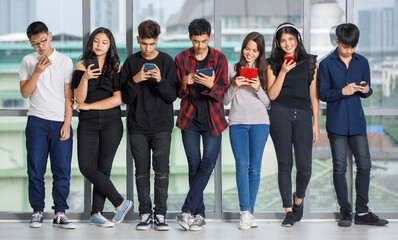 Obraz premium Group of seven young male and female teenagers in casual clothes standing and using their smartphones and tablet. College friends playing mobile phones. Concept of a phone addicted in adolescent