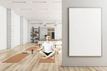  Young man in lotus pose sitting in modern yoga gym interior with empty poster on wall, equipment and daylight. Health and sport concept. Mock up. © Who is Danny