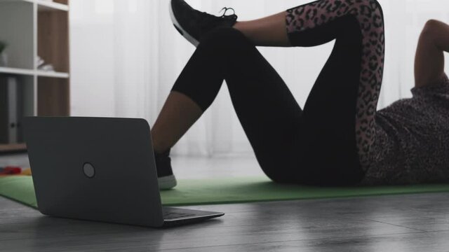 Active fitness. Sportive woman. Online training. Good body shape. Athletic lady in sportswear doing side trunk curls workout with laptop on floor in shadow light room interior.