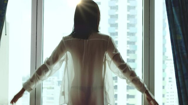 Woman in a shear shirt with the light silhouetting her perfect body opens the bedroom curtains to let in the light and greet a new day of opportunities