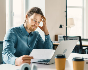 Young upset businessman looking at financial document or bill with frustrated face expression while sitting behind desk with modern laptop computer in office, entrepreneur receiving debt notification