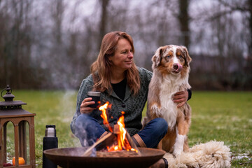 Dog sits by the fire with the hosts and HuskyAustralian Shepherd sits next to his owner by the fire. The woman drinks coffee from a thermos cup. Together they sit on a fur rug on the snow-covered