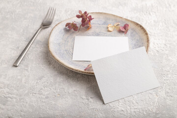 White paper invitation card, mockup with dried hydrangea flowers on ceramic plate and gray concrete background. side view, copy space.