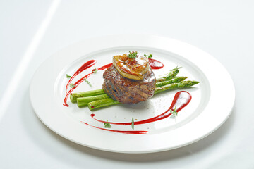 Portion of grilled mignon steak with foie gras and asparagus, berry sauce in a white plate. Isolated on grey background. - obrazy, fototapety, plakaty