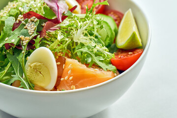 Delicious veggie bowl with salmon, avocado, cucumber, cherry tomatoes and mix salad in a white plate. Isolated on grey background.