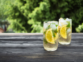 Cold carbonated lemonade with ice, lemon and currant leaves in a transparent glass. Summer drink stands on a wooden table in the garden. Background - fir branches