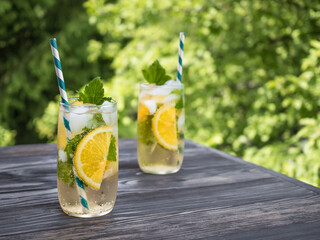 Cold carbonated lemonade with ice, lemon, currant leaves and a cocktail tube in a transparent glass. Summer drink stands on a wooden table in the garden. Background - green tree branches