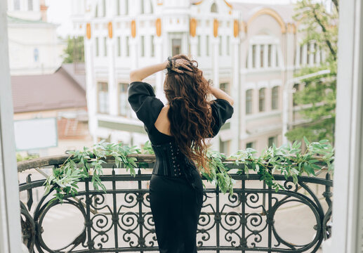 Luxury Woman In Black Evening Dress Stands On Balcony Looks At City. Back To Camera Rear View, Turned Away Face. Mysterious Silhouette Of Glamorous Girl Queen. Hands Hold Long Hair, Fly In Wind Motion