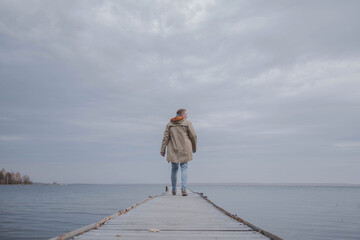 A young blond man in a light raincoat walks on a wooden pier on a lake on a foggy autumn day...