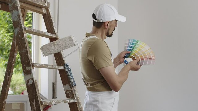 house painter man decorator choose the color using the sample swatch, work the white wall of the house to renovate, a wooden ladder, a paint roller and a green window as a background