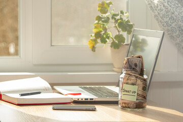 A glass jar with STARTUP written on it, with coins and banknotes inside stands on a desk.The concept is funding and promoting an innovative idea. Laptop, smartphone, diary on the desk.