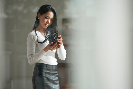 Serious vintage Asian female photographer in leather skirt analyzing photos on old-fashioned camera