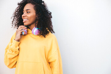 Beautiful black woman with afro curls hairstyle.Smiling model in yellow hoodie.Sexy carefree female enjoying listening music in wireless headphones.Posing on street background near wall