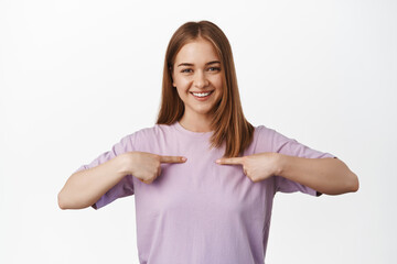 Image of blond girl in t-shirt pointing at chest, showing herself, logo on clothes, smiling happy, standing pleased against white background