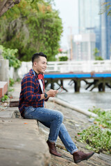 Fototapeta premium Smiling dreamy young Asian man with red headphones around neck sitting on ground near riverside and choosing track on smartphone