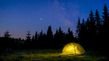 Romantic view with a tent on a meadow in the middle of a forest under a starry sky. Overnight in the mountains, in the wild during the hike.